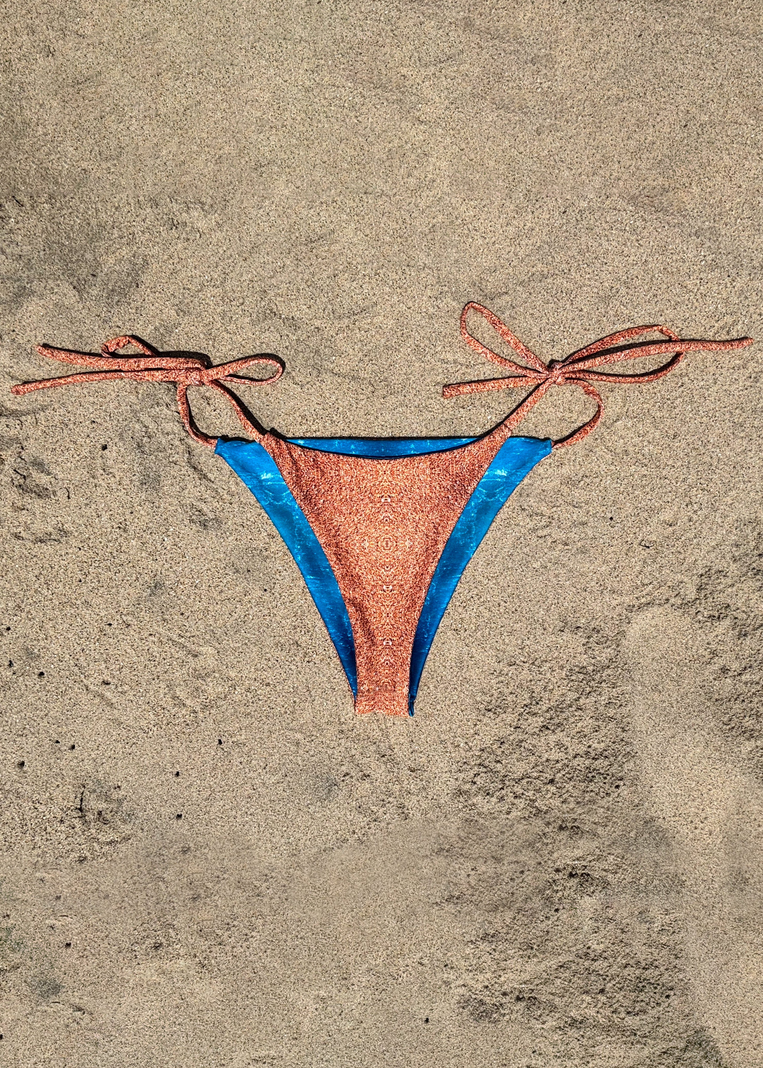 A string bikini bottom laid flat on sand, featuring a copper-orange reversible side with bright blue side panels and matching copper-orange sand print tie strings