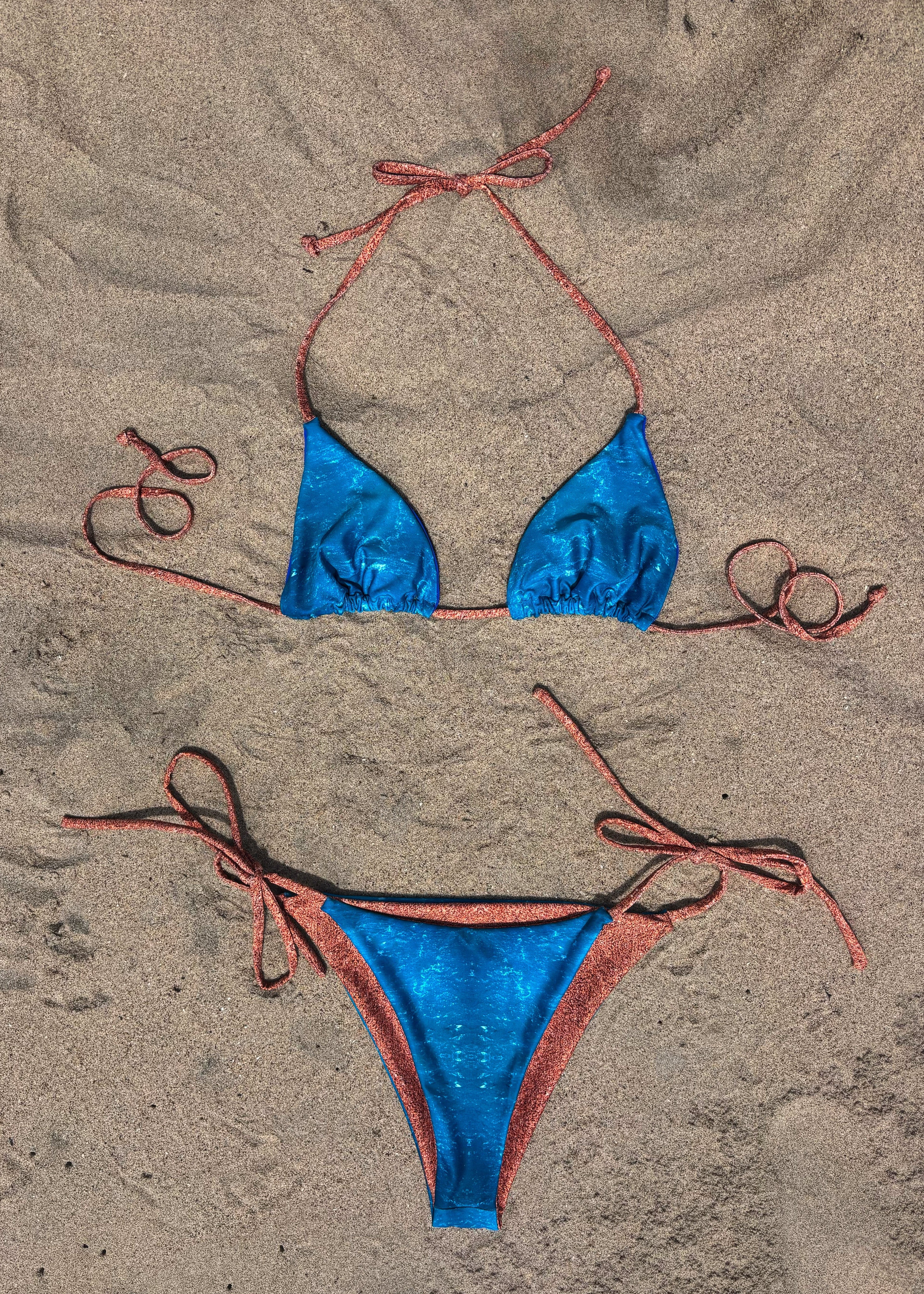 A two-piece string bikini laid flat on sand, featuring a metallic blue triangle top and matching bottoms with coral-pink tie straps.