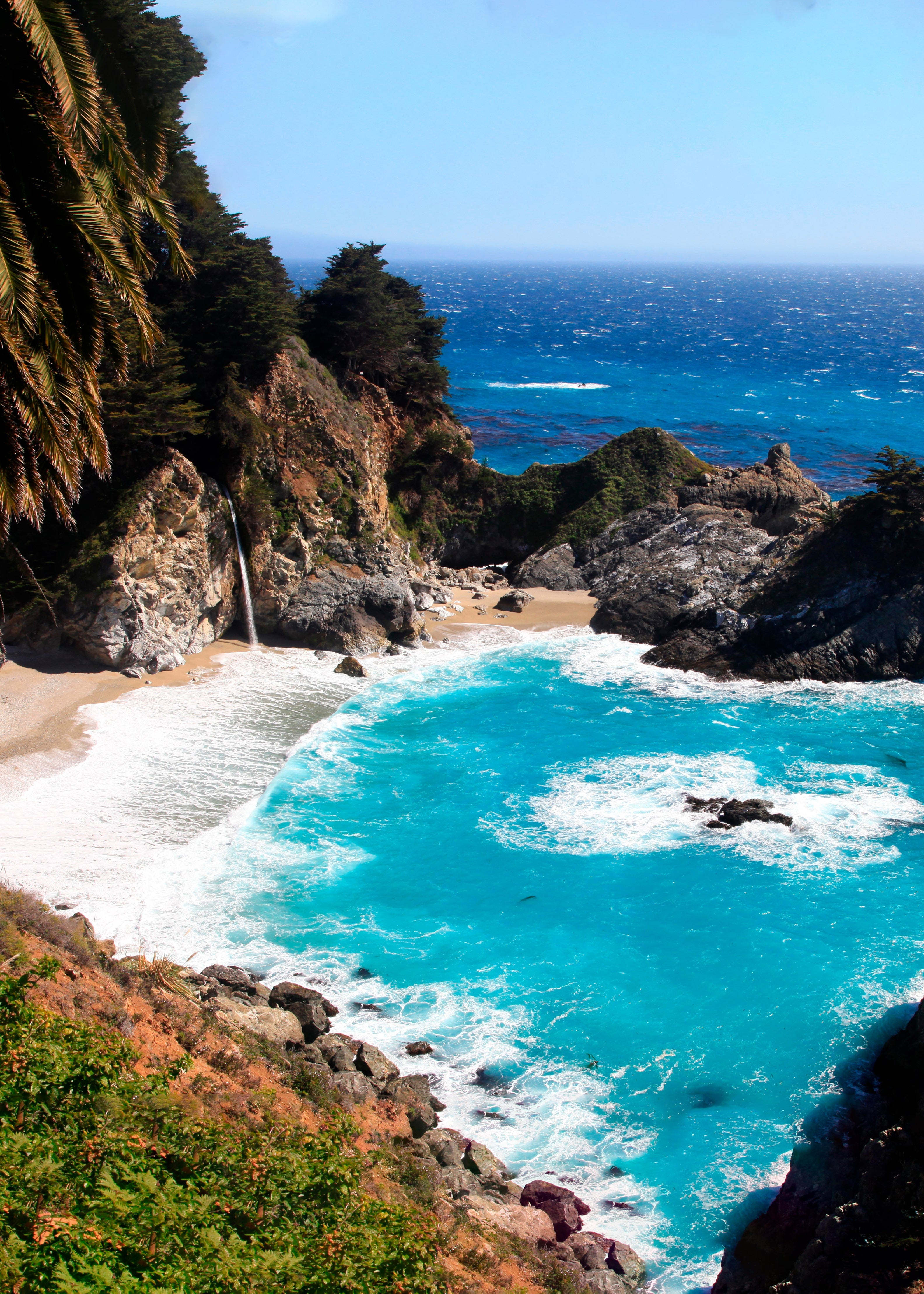 A scenic coastal cove with bright turquoise ocean water, white surf, a small sandy beach, rocky cliffs, and green trees under a clear blue sky.